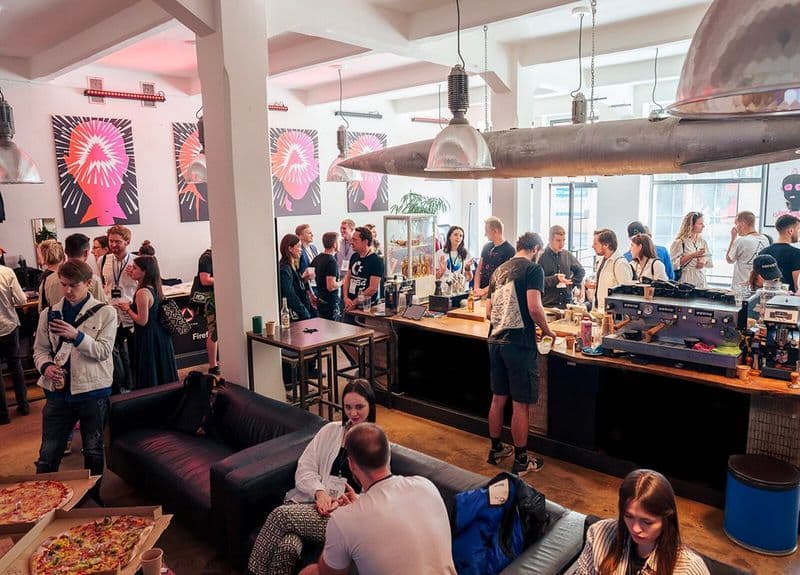 Bitcoin Coffee bar during Hackers Congress Paralelní Polis — attendees with badges at the espresso machine, red-and-pink posters on the walls, pizza boxes in the foreground.